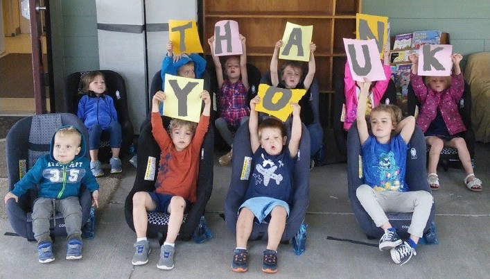 10 young children sit in car seats, 8 of them hold up papers that spell out the words 