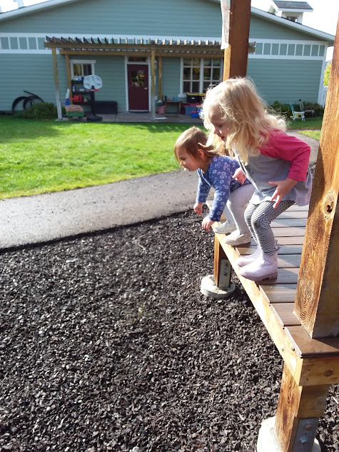 Two girls stand on a wooden play platform with knees bent, preparing to jump onto the woodchips below. In the background is a single-story school building.